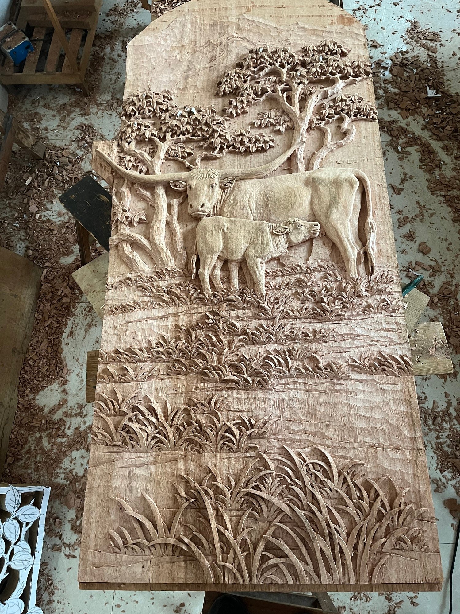 Overhead view of a full arched solid wood door panel resting on a workshop workbench surrounded by wood shavings, featuring hand-carved Texas Longhorn cattle relief art for a rustic ranch home.