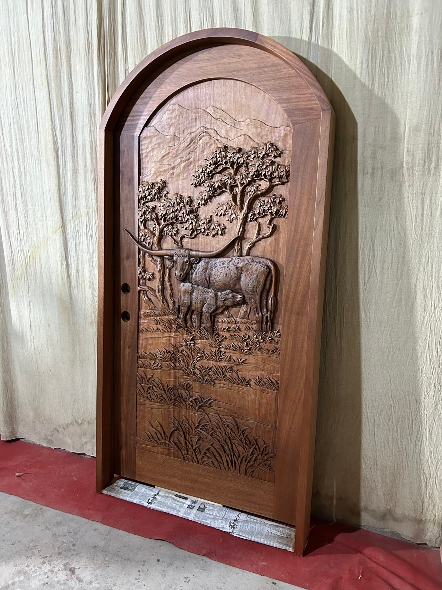 Angled three-quarter vertical view of a finished round-top arched solid wood single entry door mounted inside its matching door jamb frame, featuring a hand-carved Texas Longhorn cow and calf relief for a Texas ranch home.