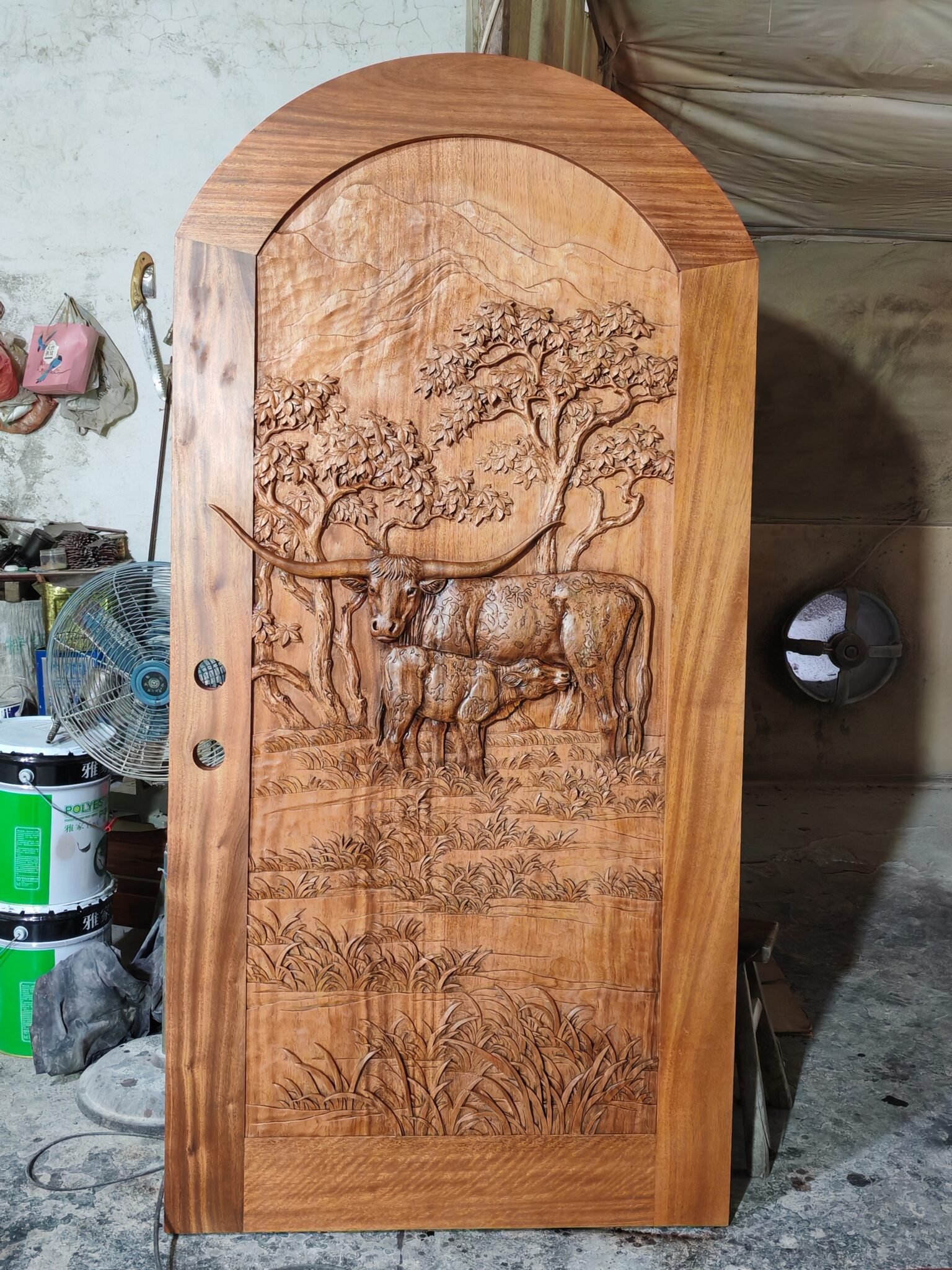 Frontal full-length view of a completed arched solid wood single entry door with clear protective topcoat standing in a workshop, featuring a Texas Longhorn cow and calf relief design for a Texas ranch home.