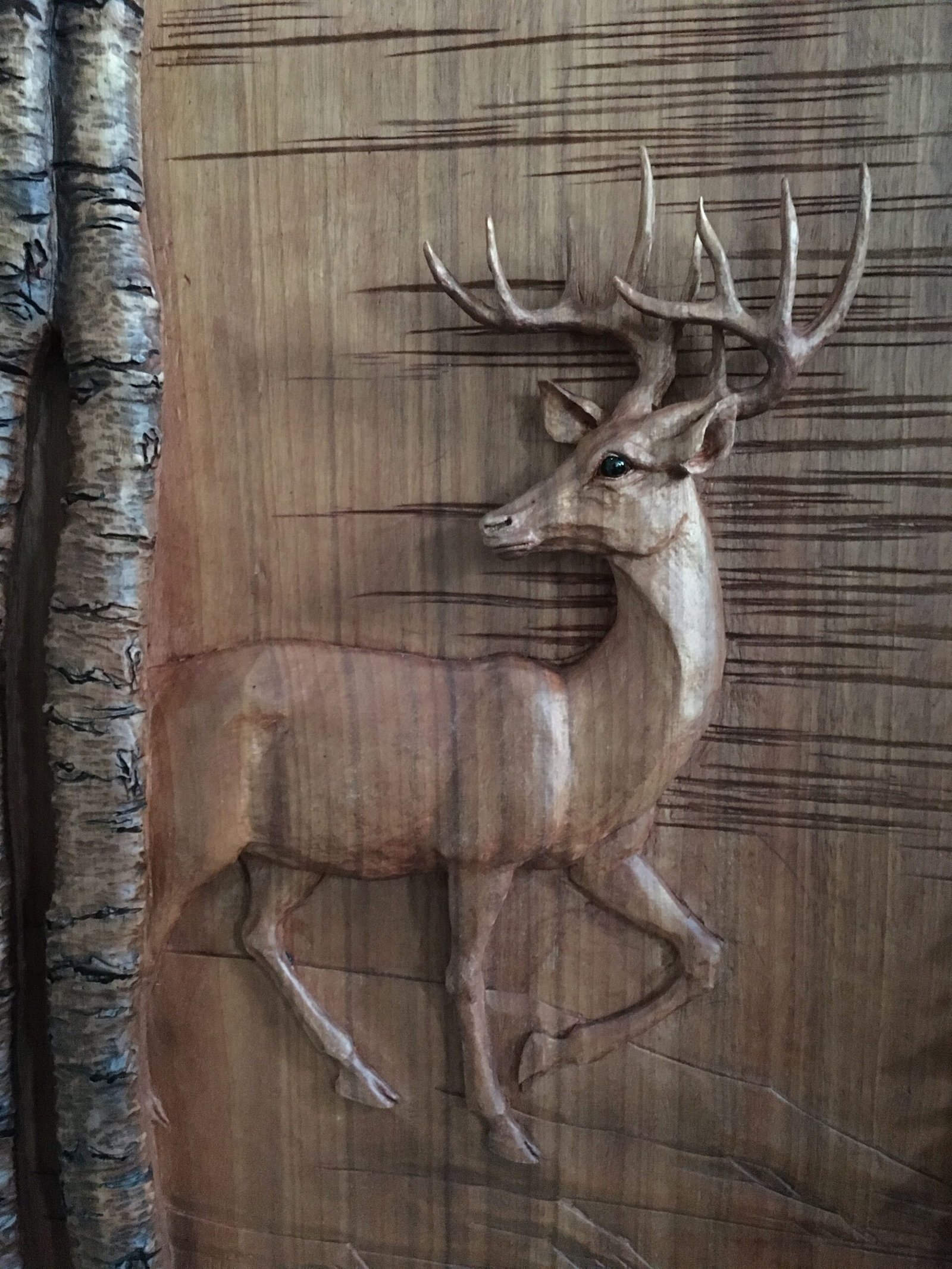 Close-up of a lifelike hand-carved wooden deer (stag with full antlers) in high-relief on a solid wood custom door panel, shown next to textured birch tree bark, showcasing intricate craftsmanship by Crown Custom Doors