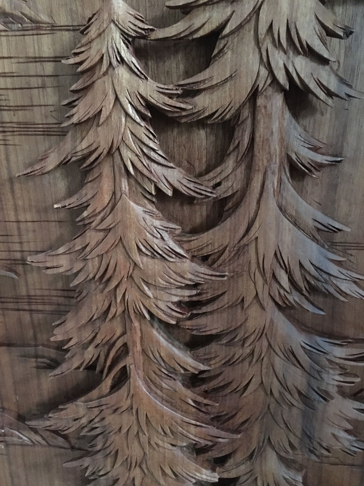 Close-up of intricately hand-carved wooden pine trees with layered needle-like branches in high-relief on a solid wood custom door panel, showcasing detailed craftsmanship by Crown Custom Doors