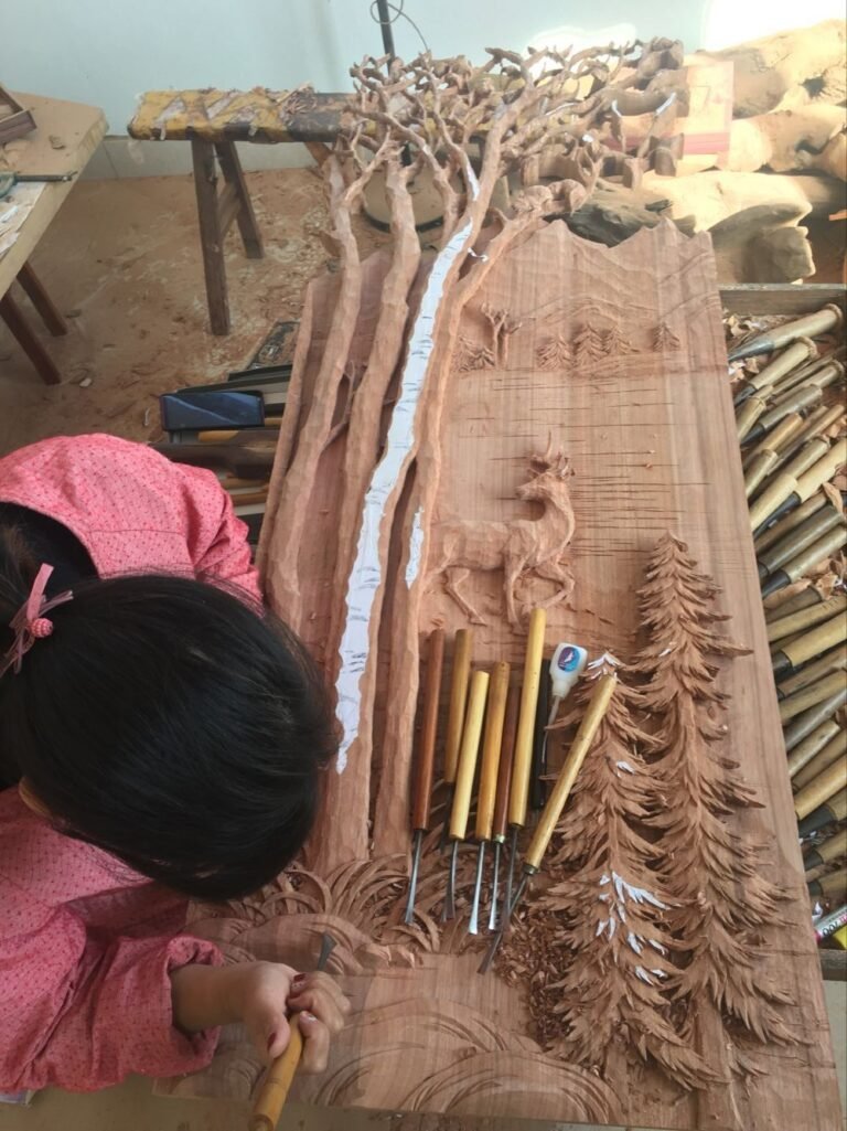 Artisan hand-carving a large solid wood panel for a custom entry door featuring aspen trees, deer, pine trees, mountains and lake scene; close-up of detailed high-relief work with carving tools and wood shavings in the Crown Custom Doors workshop