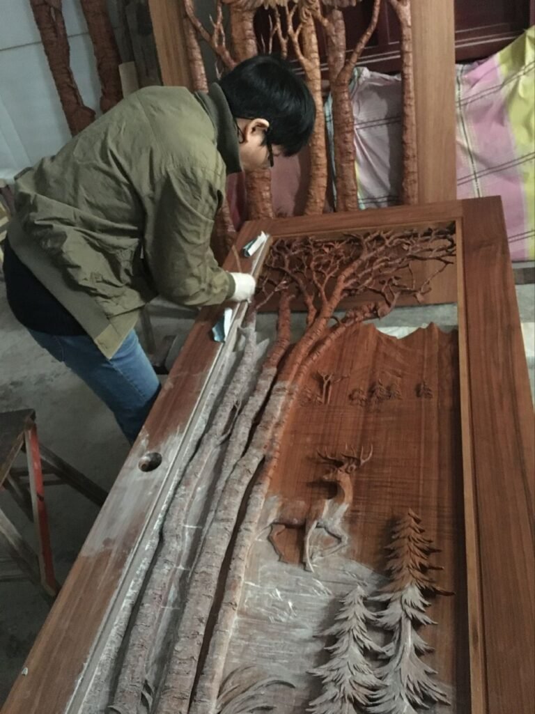 Artisan in workshop carefully hand-finishing a large solid wood custom door panel featuring high-relief aspen trees, deer, pine trees, mountains and lake scene; white gloves and tools visible during the detailed refinement process by Crown Custom Doors