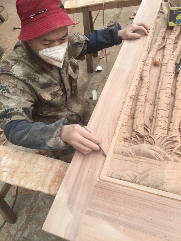Skilled craftsman wearing a red hat and mask carefully hand-sanding the edge of a large solid wood custom door panel with high-relief tree and foliage carvings before staining, shown in the Crown Custom Doors workshop
