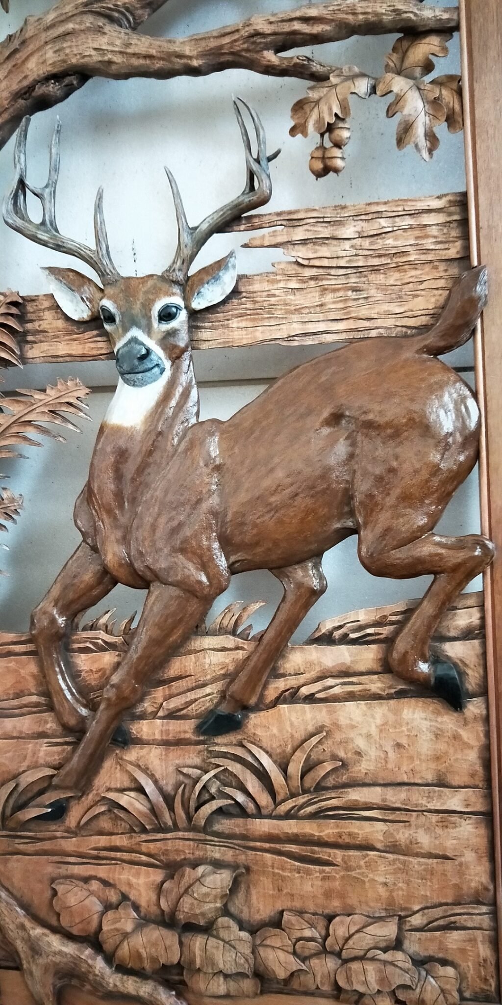 Close-up shot of a solid wood door panel featuring a hand-carved and hand-painted white-tailed deer buck in mid-stride, framed by oak tree branches, acorns, ferns, and forest floor foliage over a glass background.