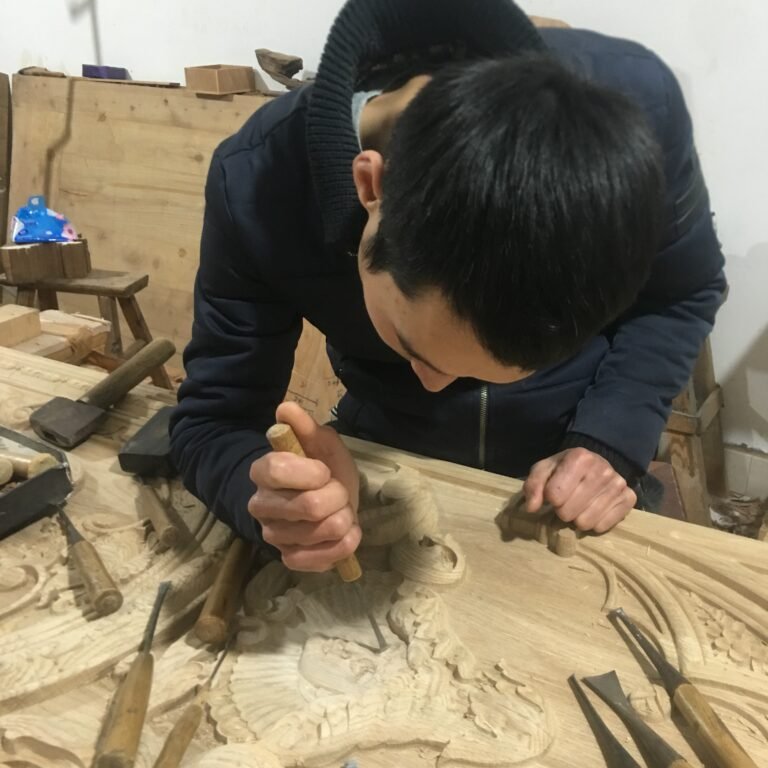 Overhead angle of a skilled woodcarver intently hand-chiseling fine relief facial features and ornamental scrollwork on a raw solid wood door panel at Crown Custom Doors.