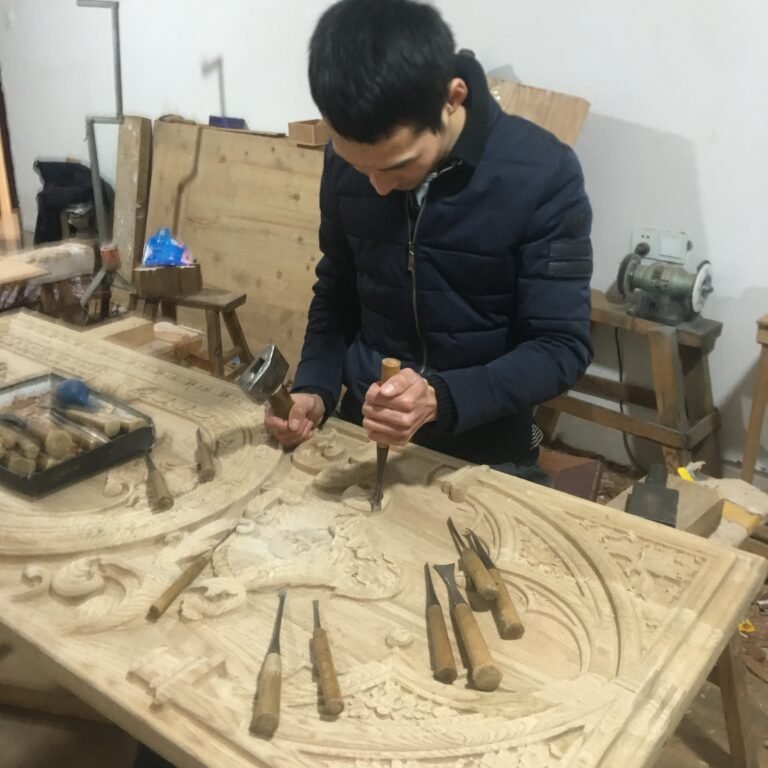 Wide workshop shot of a master craftsman using a wooden mallet and chisel to hand-carve intricate Gothic relief patterns into a large solid wood door panel surrounded by traditional woodworking tools at Crown Custom Doors.