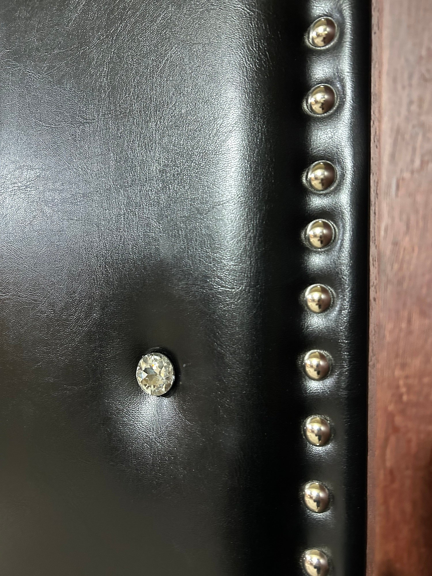 Macro shot showing fine black leather upholstery on an acoustic home theater door featuring deep diamond tufting with a faceted crystal button, hand-tacked silver metal nailhead trim, and a solid wood frame.