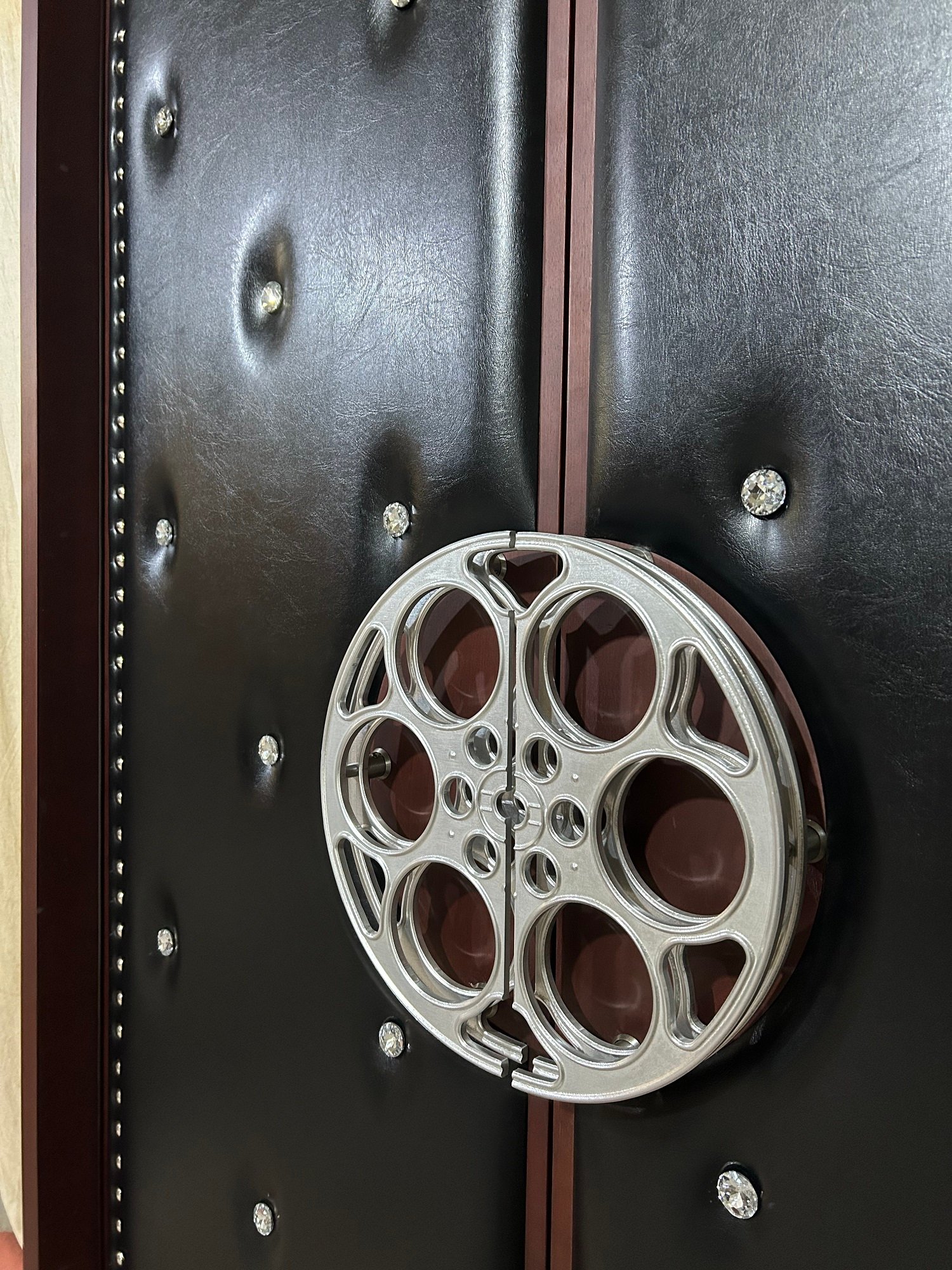 Angled close-up of custom acoustic home theater double doors showing black leather diamond tufting, crystal buttons, silver perimeter nailheads, and a silver film reel split handle set against dark reddish-brown wood jamb framing.