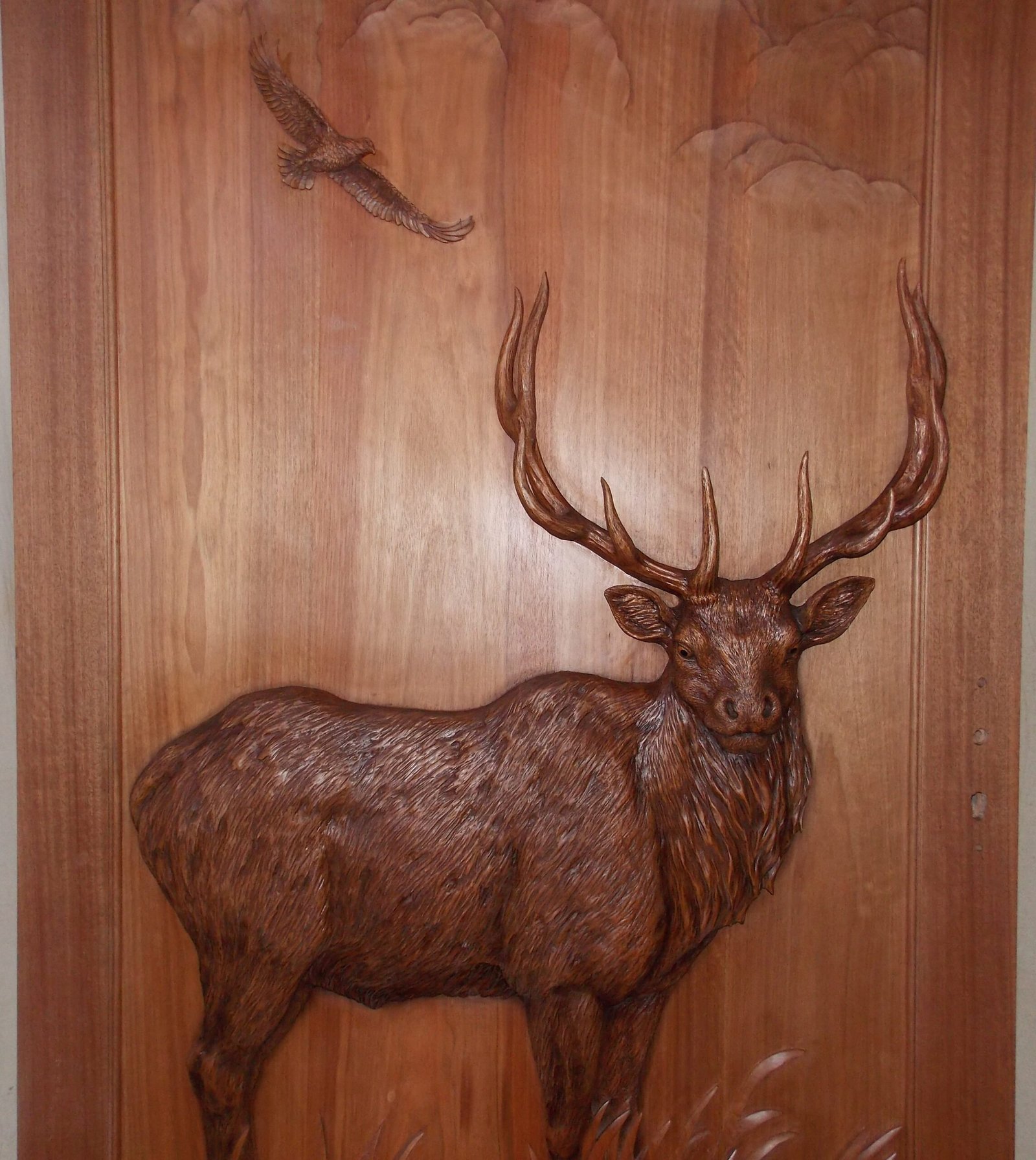 Close-up detail shot of a solid wood door panel featuring a high-relief hand-carving of a mountain stag elk with sweeping antlers set beneath a soaring eagle and carved cloud relief.