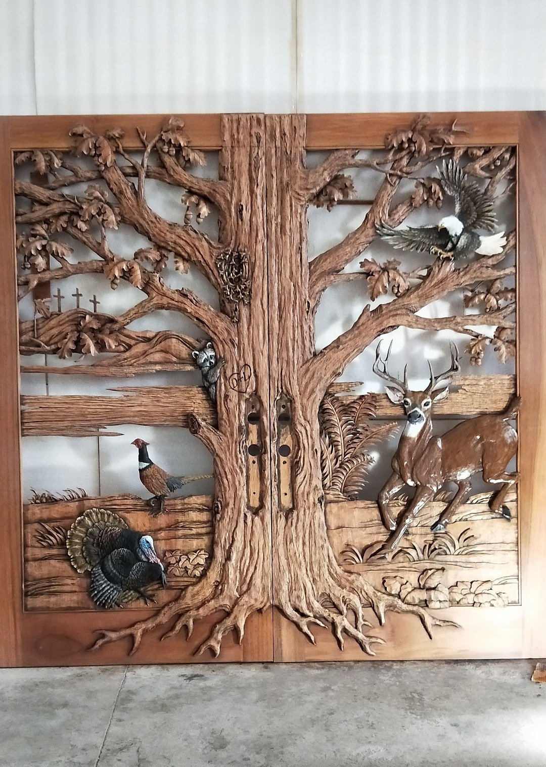 Full-length workshop view of The Nature's Embrace solid wood double entry doors, featuring a central carved oak tree trunk with spreading canopy branches, deer, wild turkey, pheasant, eagle, raccoon, bee hive, and three background crosses.