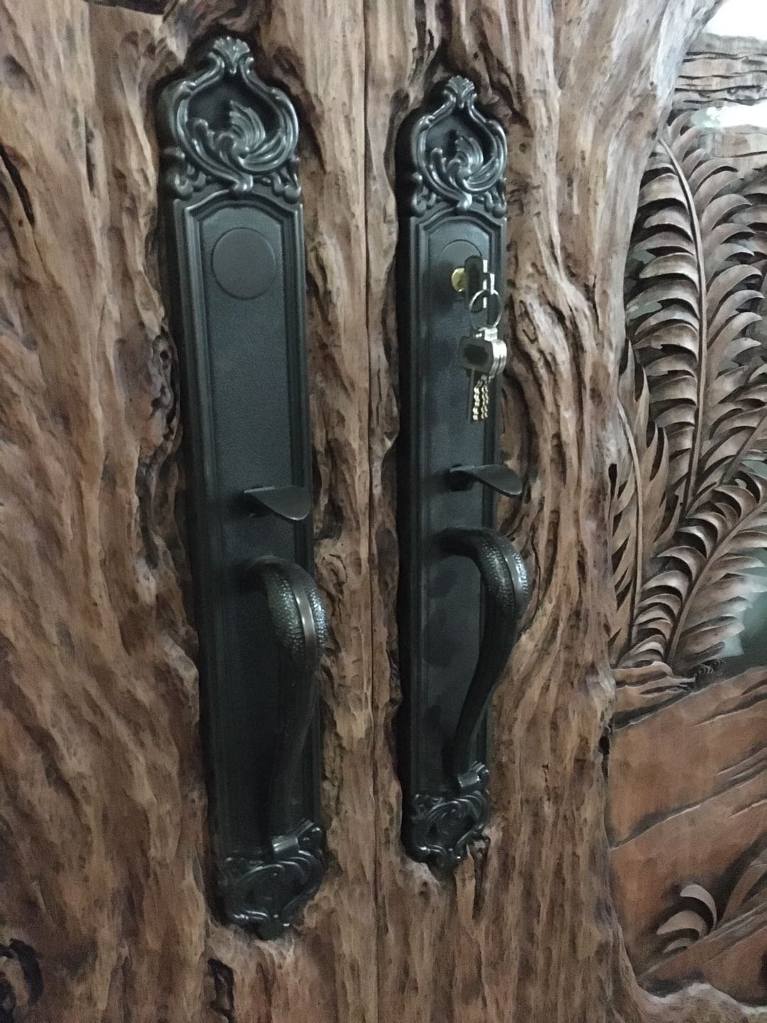 Close-up shot of dual oil-rubbed bronze decorative escutcheon handlesets with keys installed directly onto the deeply carved tree trunk bark texture of a solid wood double entry door.