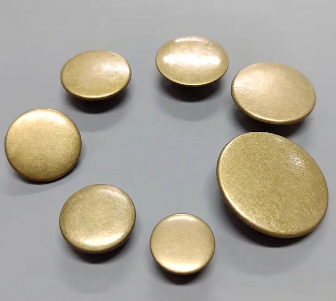 Assorted sizes of smooth, flat round solid brass decorative upholstery buttons and studs with an antique satin finish, laid out on a neutral background for custom upholstered door hardware.