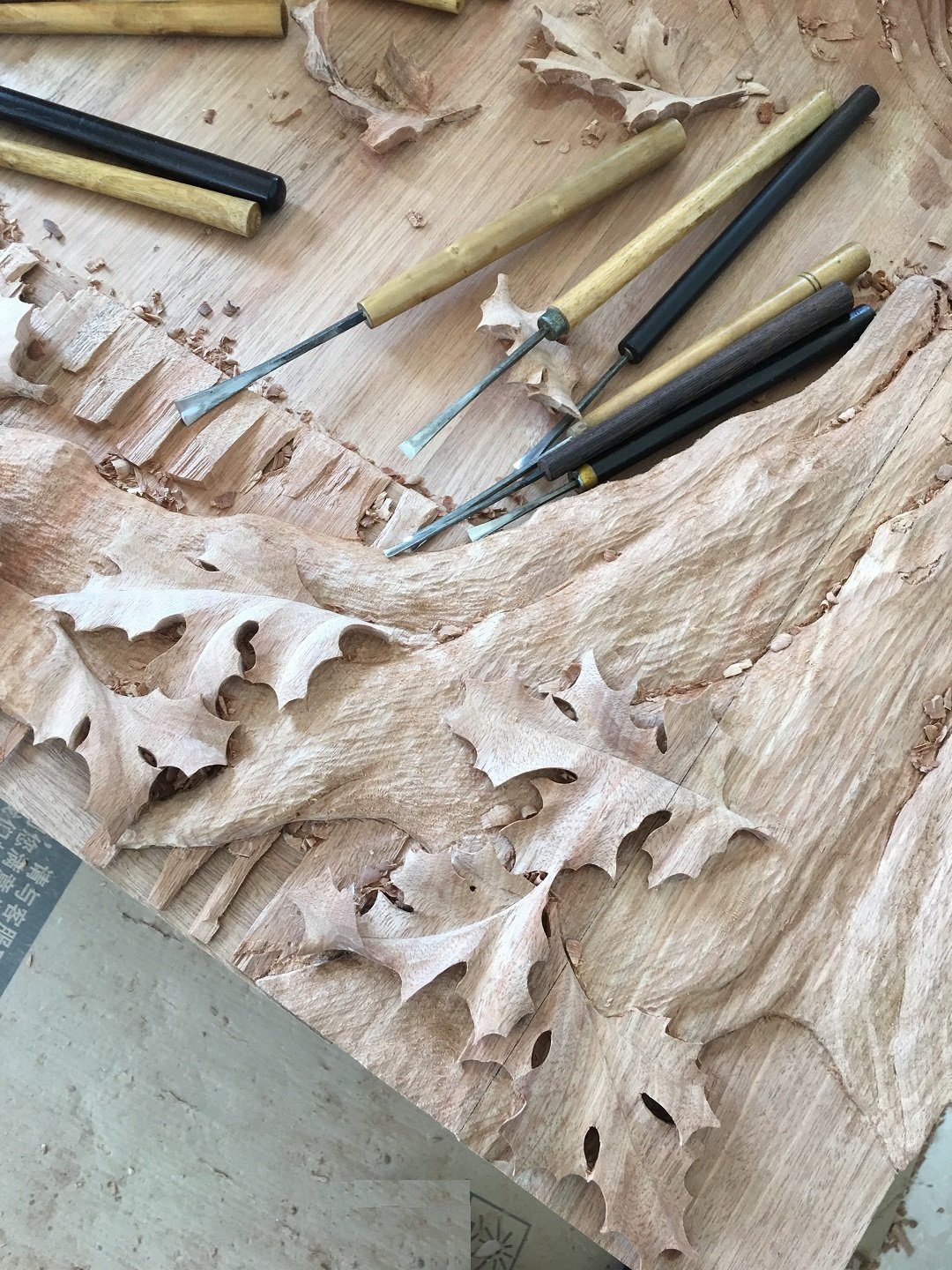 Close-up of traditional hand-carving chisels and gouges resting on raw timber next to deeply sculpted 3D oak leaf carvings and bark textures in the Crown Custom Doors workshop.