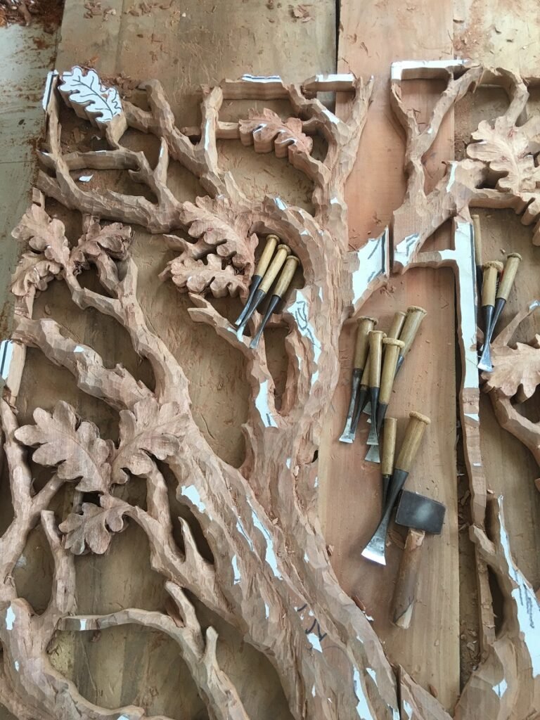 Traditional wood chisels, gouges, and small mallet resting on a solid wood door panel being hand-carved with an intricate oak tree relief pattern.
