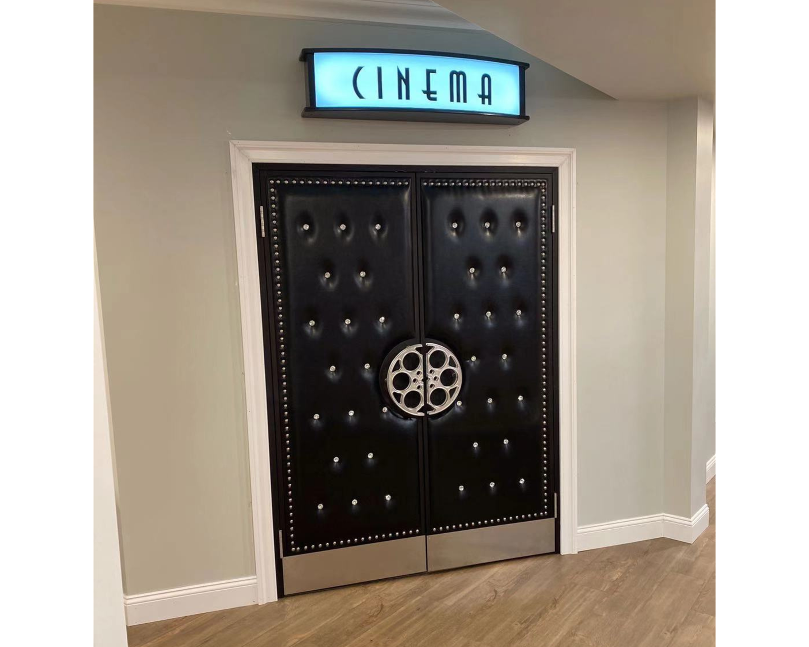 Installed custom black button-tufted vinyl acoustic home theater double doors featuring silver nailhead trim, circular silver film reel center handles, silver kickplates, and an illuminated CINEMA sign mounted above the frame.