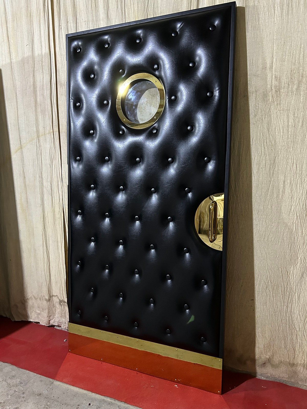 Angled side view of a custom soundproof black vinyl tufted home theater door showing deep diamond cushioning, polished brass porthole window, half-moon film reel handle, and brass kickplate.