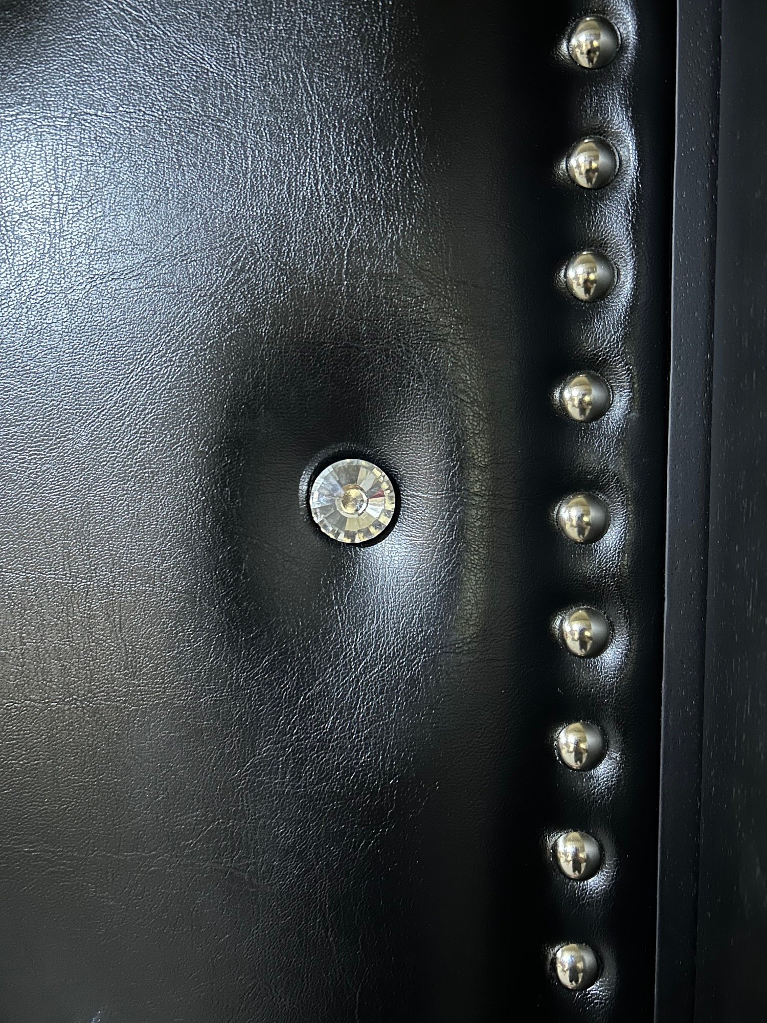 Macro close-up view of black leatherette upholstery showing textured vinyl grain, deep-set faceted crystal diamond tufting button, and a row of polished silver studs along the edge frame of a custom acoustic home theater door.