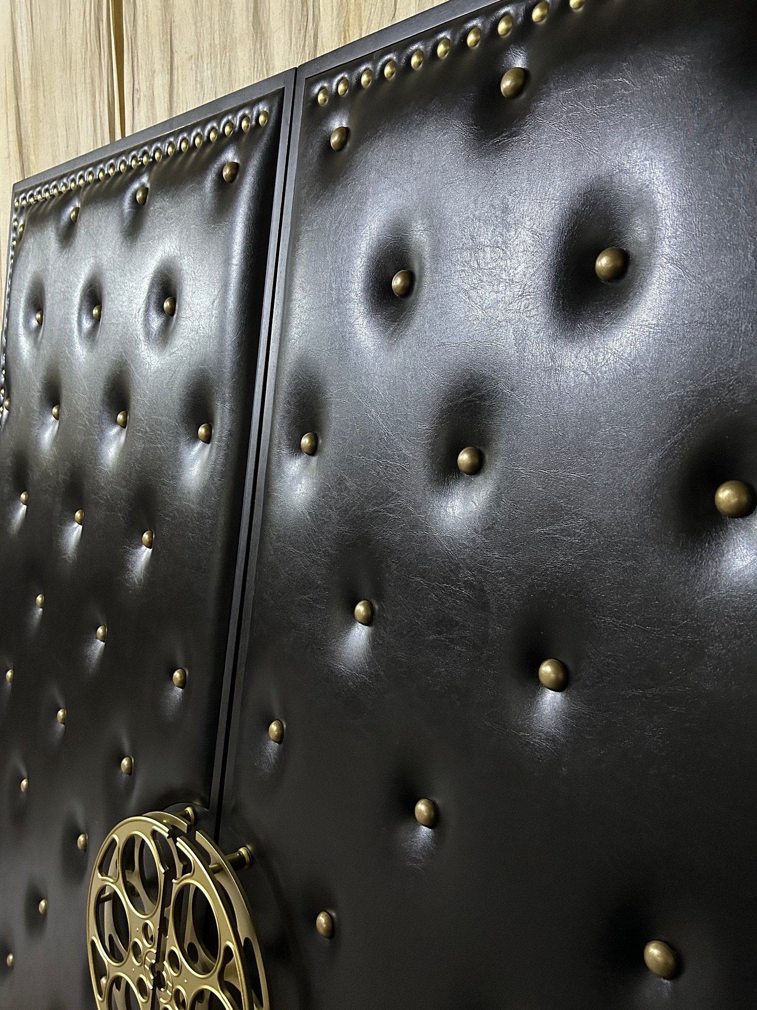 Deep side-angle view of custom black leather-look vinyl upholstered double doors featuring diamond tufting, antique brass nailhead studs, and a brass film reel handle at the bottom.
