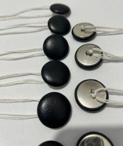 Close-up view of black leatherette covered upholstery buttons displaying both textured front faces and metal wire shank backs threaded with heavy-duty white tufting twine.