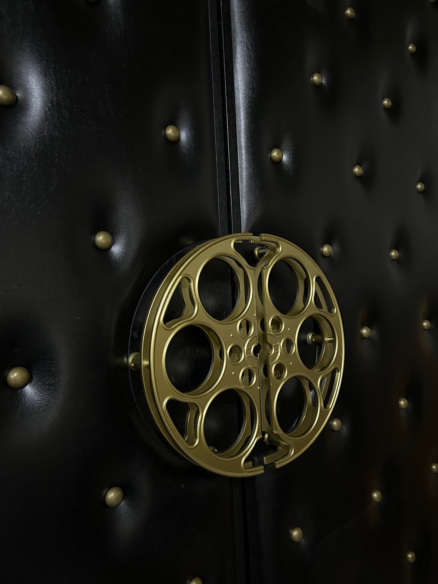 Angled side view showing the dimensional depth of custom brass film reel door handles mounted on black tufted vinyl upholstery with antique brass studs.