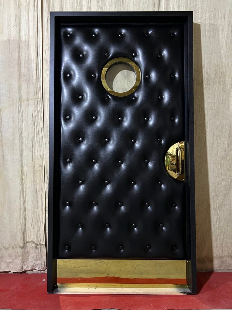 Custom acoustic home theater door upholstered in black button-tufted vinyl featuring a circular brass porthole window, brass half-moon pull handle, and polished brass kickplate within a black jamb frame.