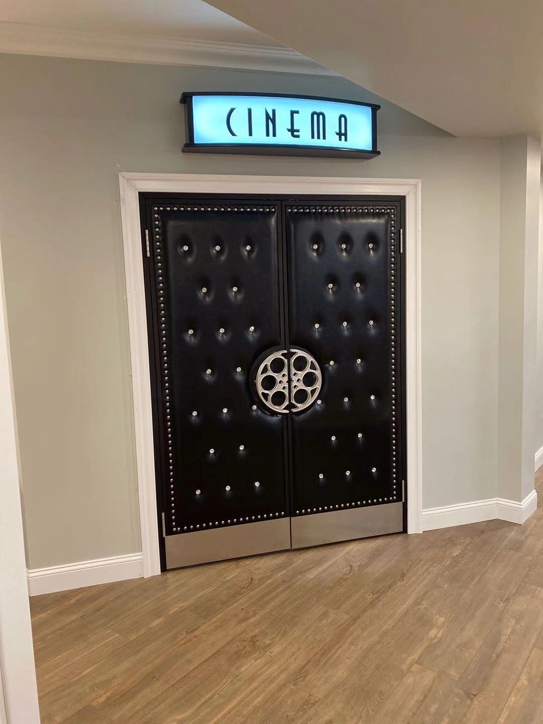 Installed custom black button-tufted vinyl acoustic home theater double doors featuring silver nailhead trim, a circular silver film reel center pull handle, stainless kickplates, and a blue backlit CINEMA marquee sign overhead.