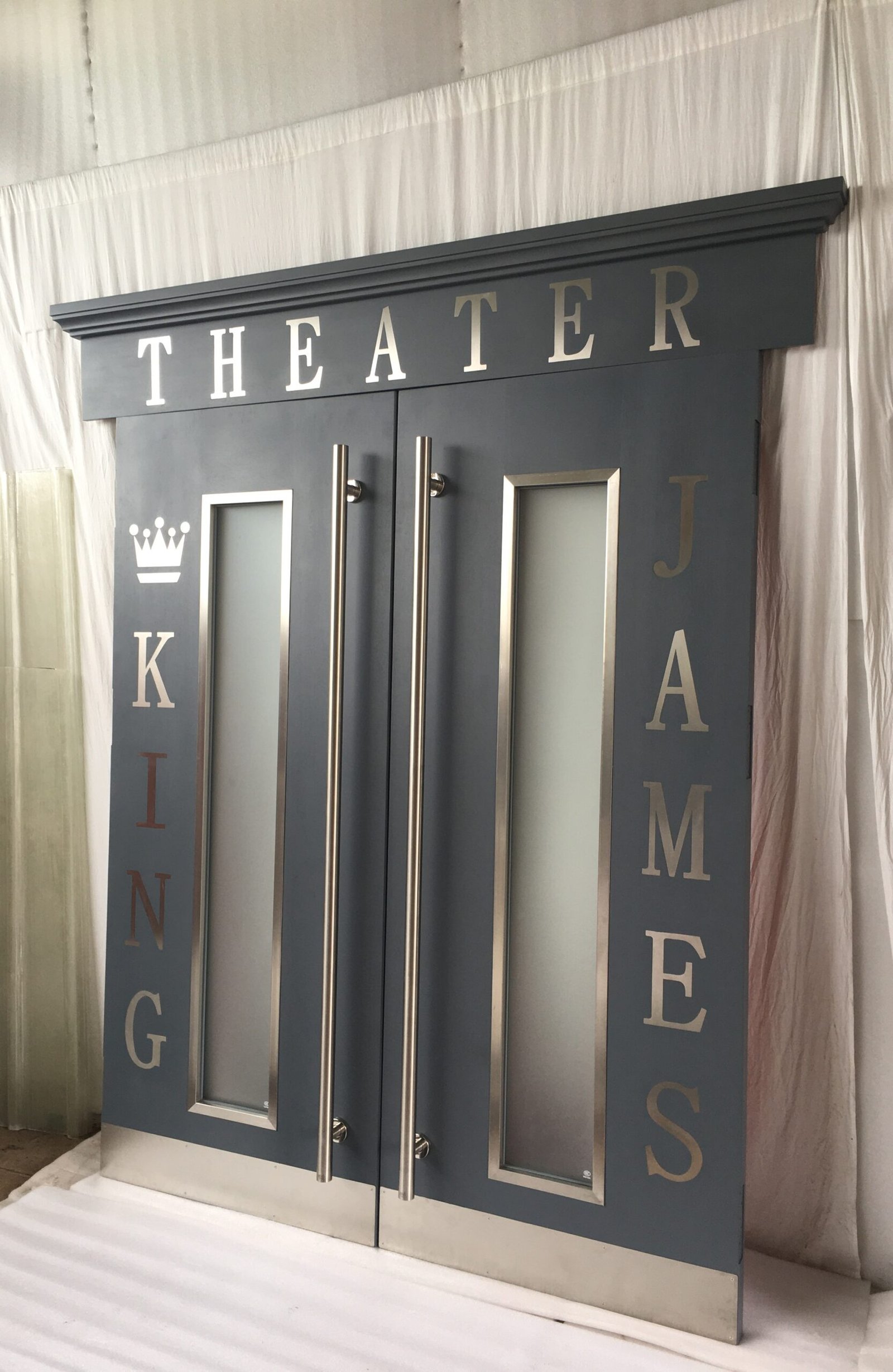 Angled studio shot of custom charcoal gray home theater double doors featuring long frosted vision glass, tall brushed stainless steel pull handles, gold "King James Theater" lettering, and crown header molding by Crown Custom Doors.