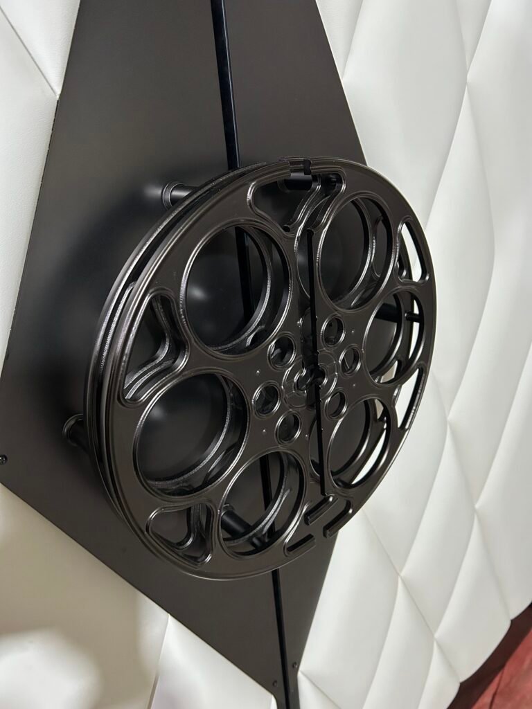 Angled close-up view of a custom-crafted matte black aluminum film-reel door pull handle mounted on diamond stainless steel push plates over off-white diamond-tufted upholstered theater doors.