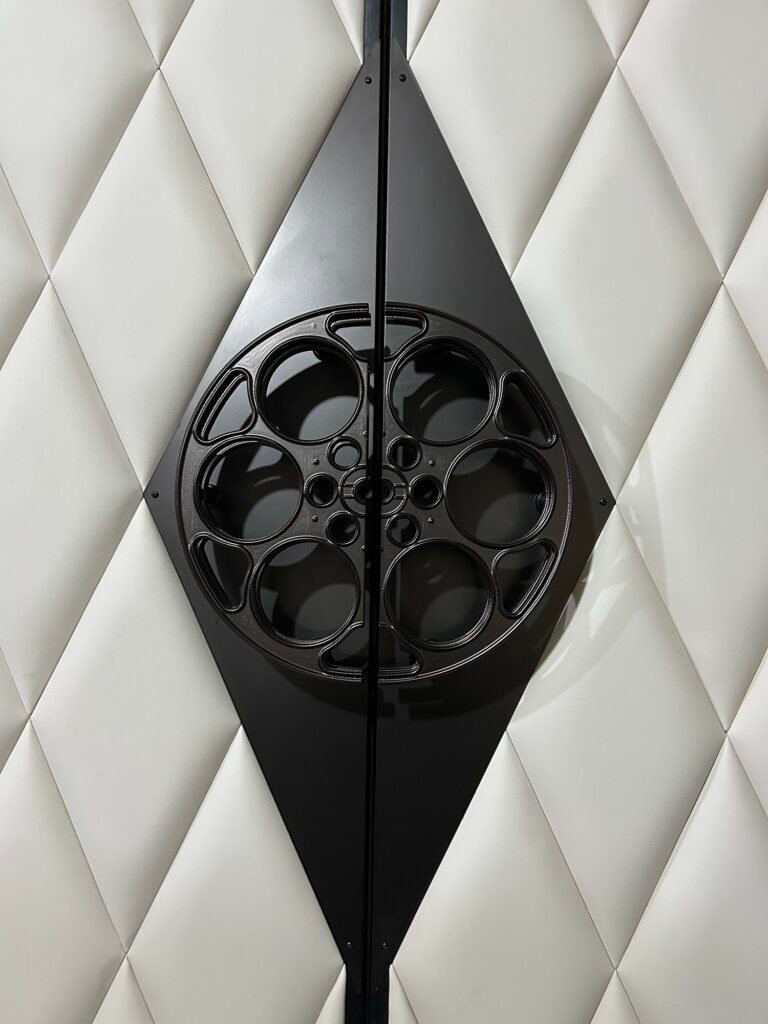 High-resolution vertical close-up of a custom matte black film-reel door pull handle and integrated diamond push plates mounted on off-white diamond-tufted vinyl upholstered double doors.