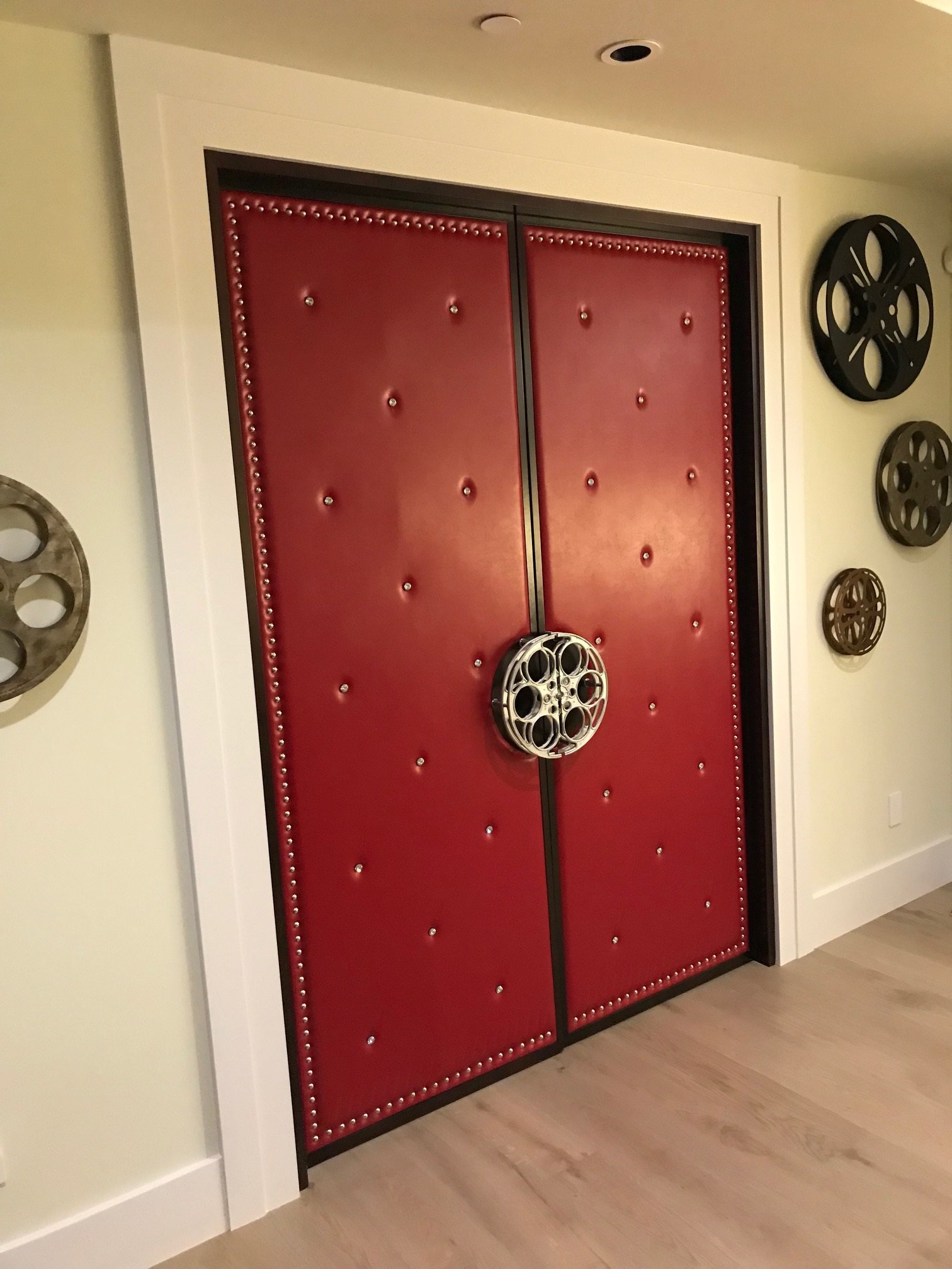 Custom red upholstered soundproof home theater double doors with diamond-tufted accents, silver decorative stud trim, and central movie film reel door handles.