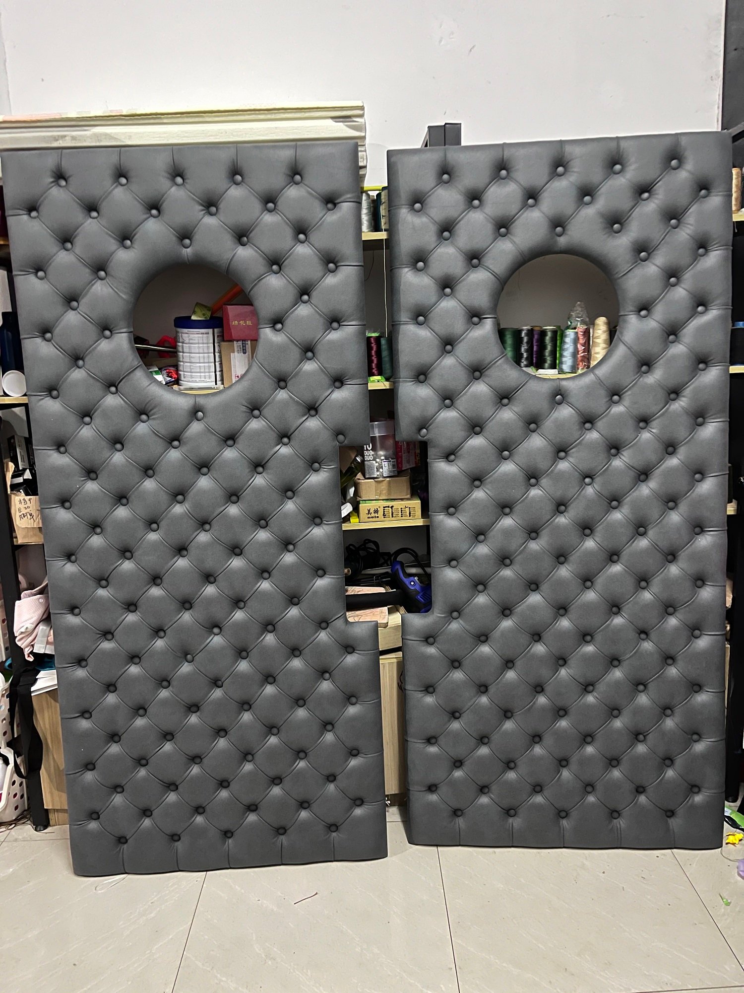 Pair of handcrafted charcoal grey diamond-tufted upholstery panels with circular porthole cutouts and pull-handle notches in workshop production for a custom acoustic theater door.
