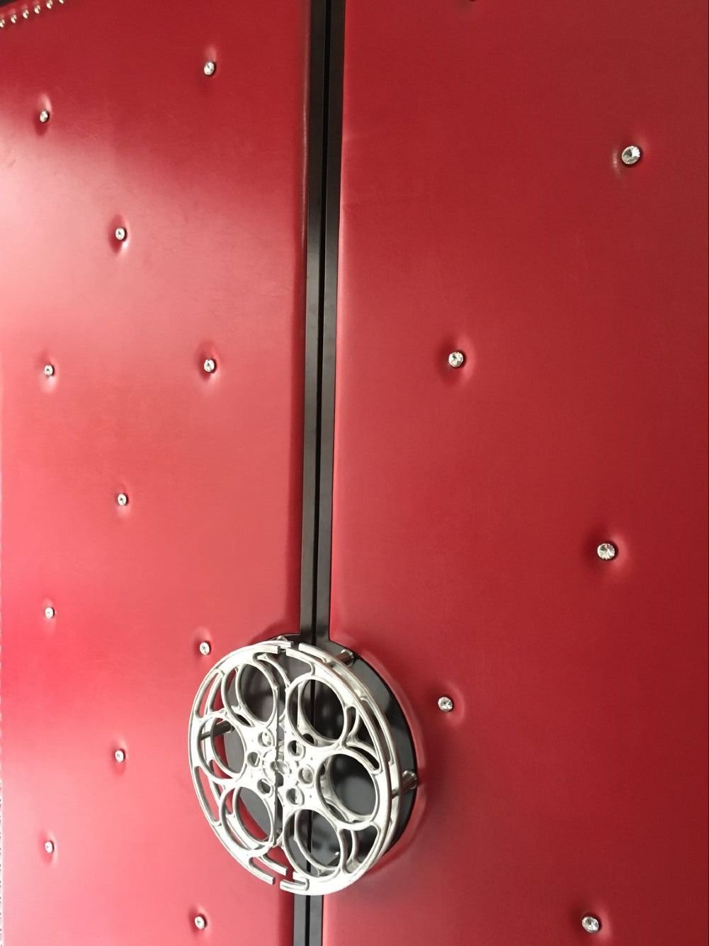 Close-up detail of a metallic movie film reel handle on custom red vinyl upholstered double doors with diamond-tufted crystal buttons.