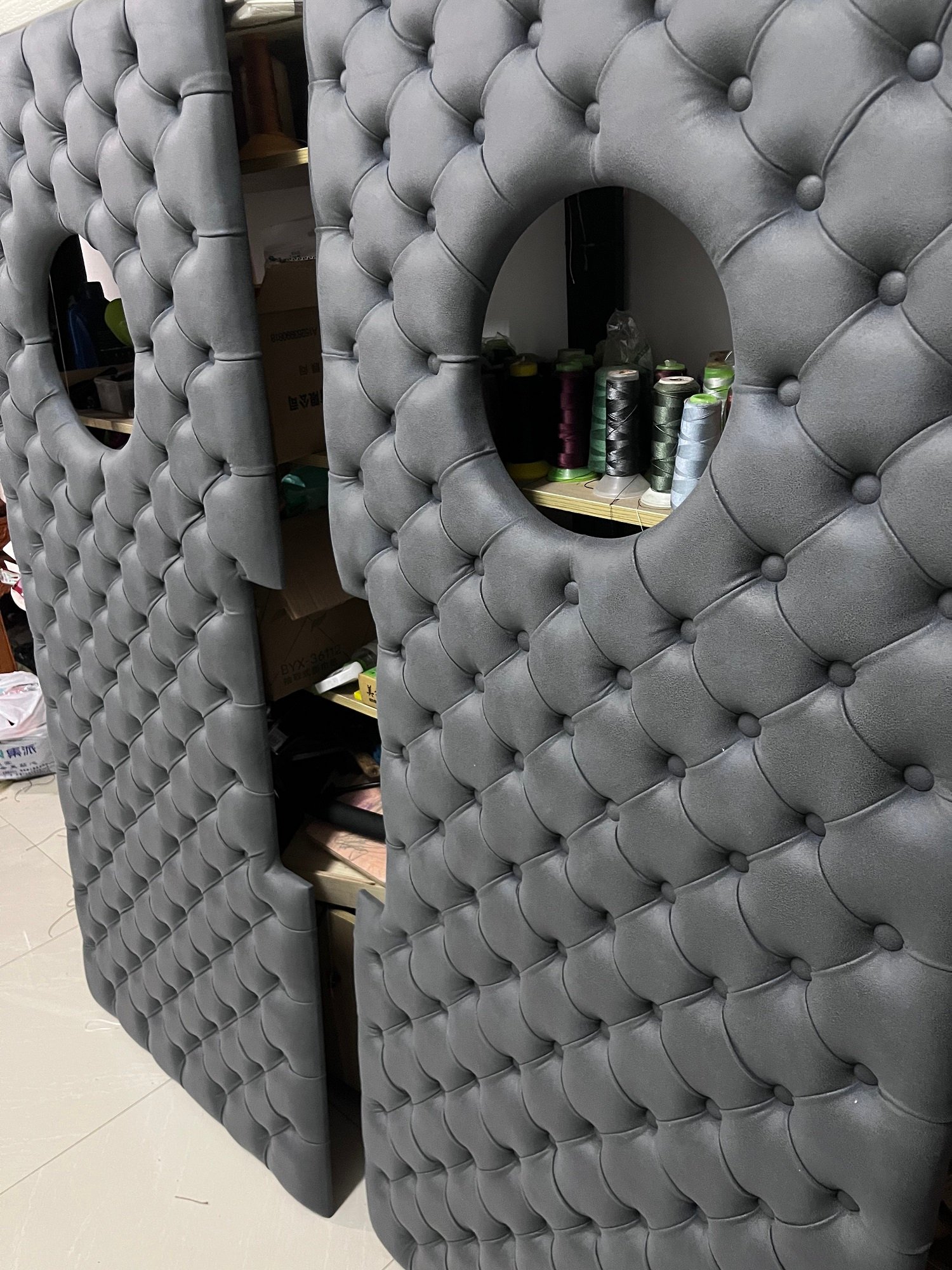 Close-up angle of handcrafted charcoal grey diamond-tufted vinyl upholstery panels showing detailed button-tacking, porthole cutouts, and side handle notches in a workshop setting.