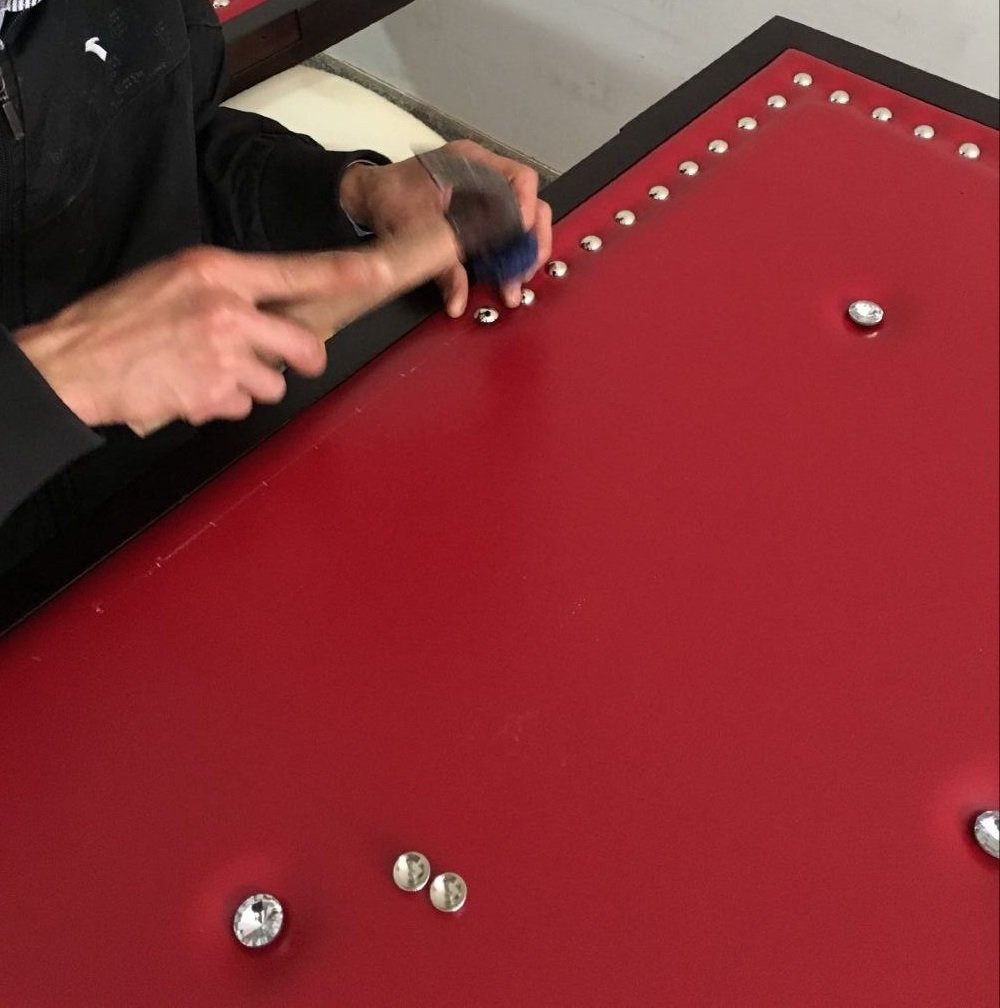 Wood artisan hand-tacking silver decorative studs and crystal tufting buttons onto a custom red vinyl upholstered home theater door panel.