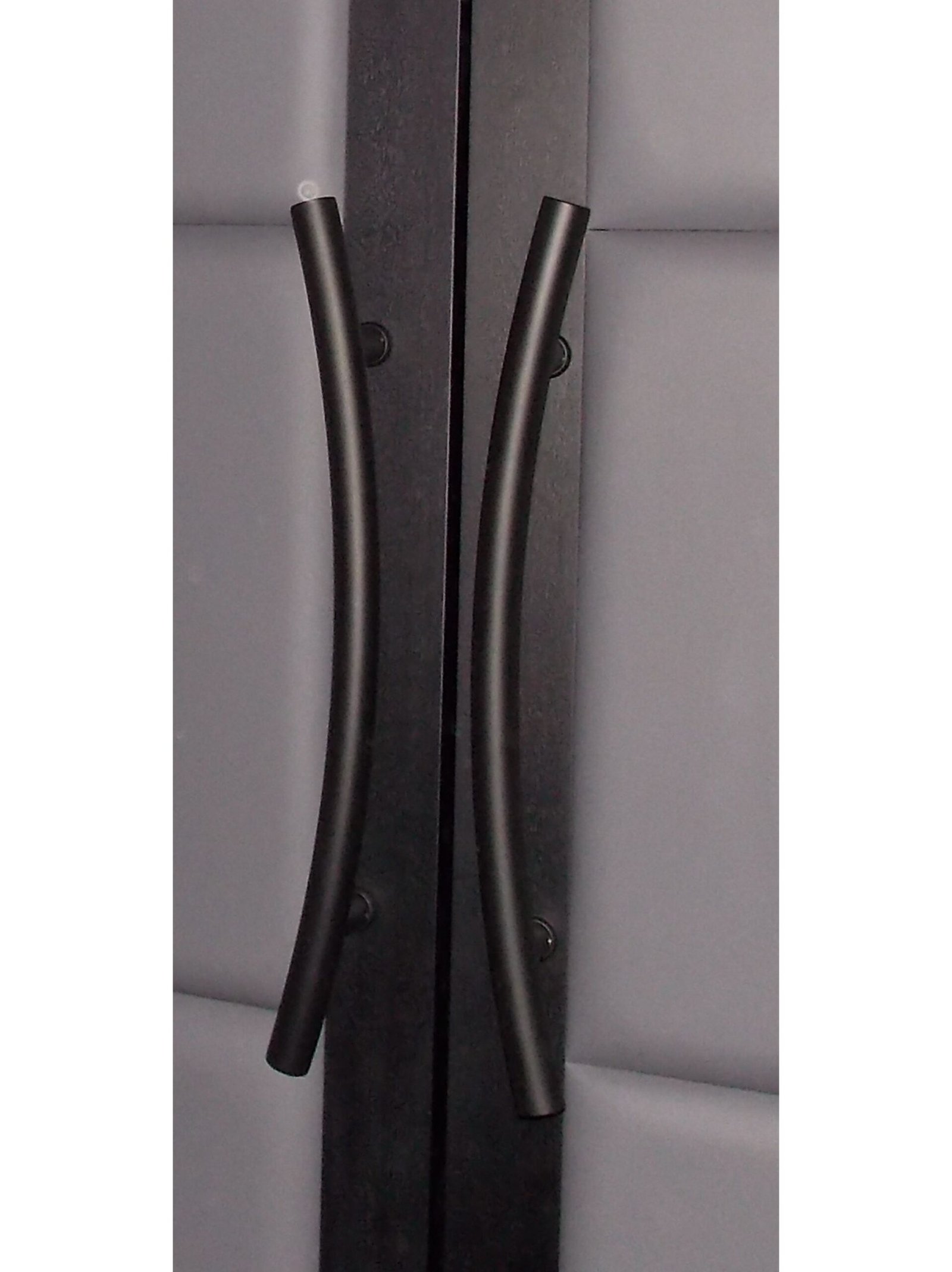 Close-up view of sleek curved matte black metal pull handles on dark wood double doors with storm gray padded leather panels by Crown Custom Doors.