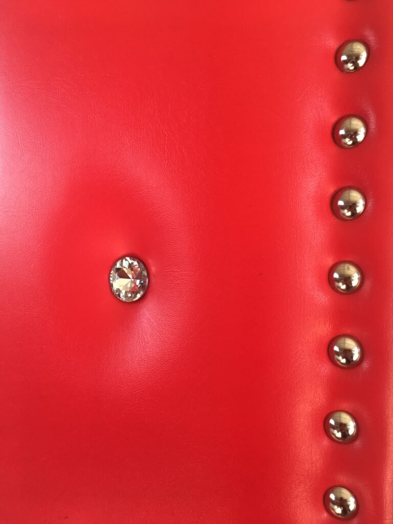 Extreme close-up macro view of vibrant red vinyl leatherette upholstered acoustic door padding featuring a faceted crystal button tuft and chrome polished nailhead studs by Crown Custom Doors.
