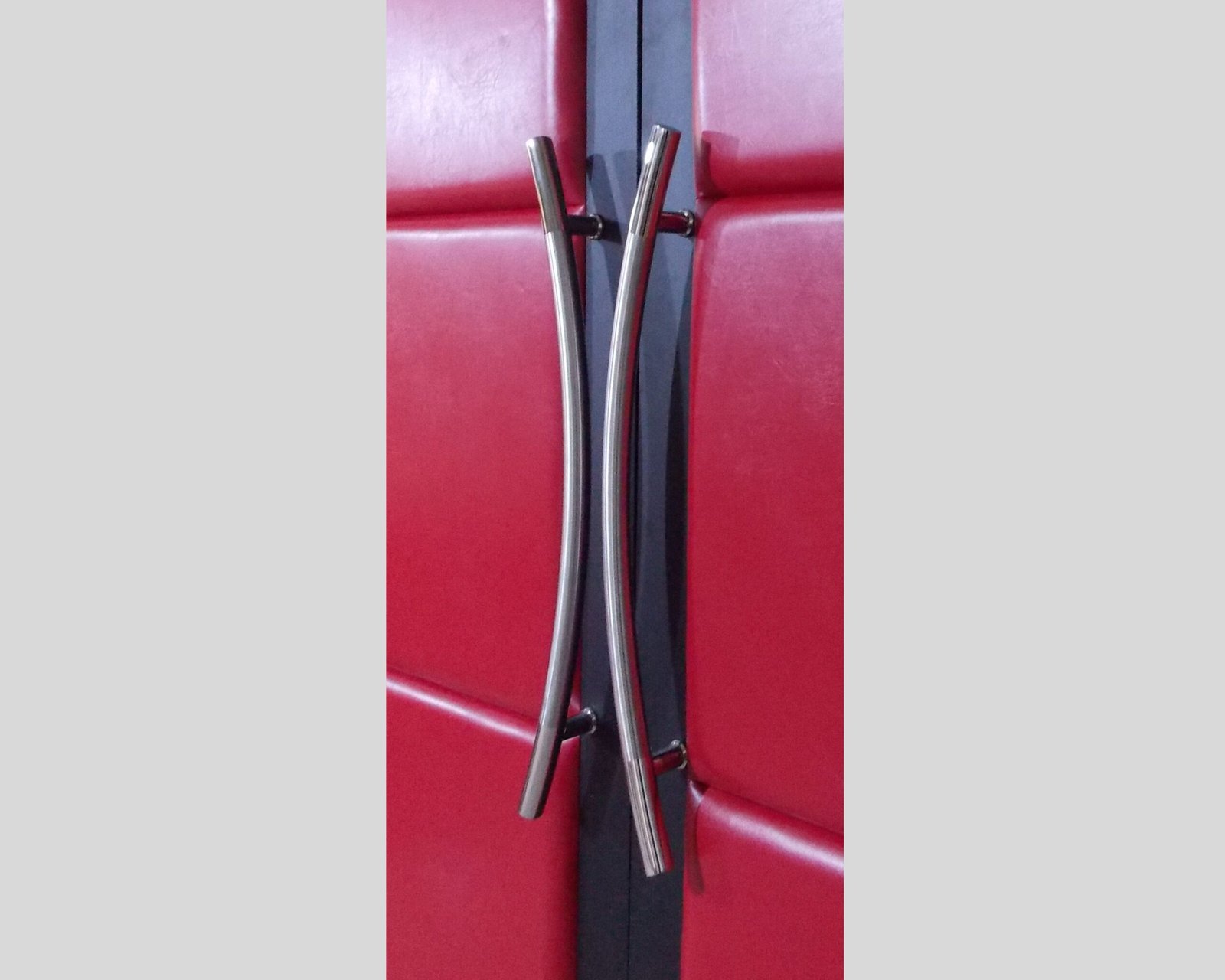 Close-up detail of dual brushed stainless steel curved ladder pull handles mounted on red padded vinyl upholstered acoustic home theater double doors by CROWNCUSTOMDOORS.