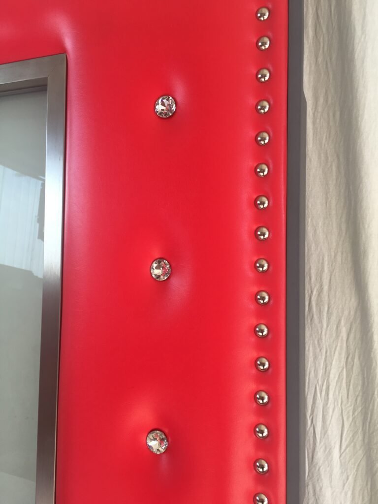 Macro close-up showing custom red vinyl leatherette upholstered acoustic door panel with diamond crystal buttons, chrome perimeter tack studs, and stainless steel glass frame trim by Crown Custom Doors.