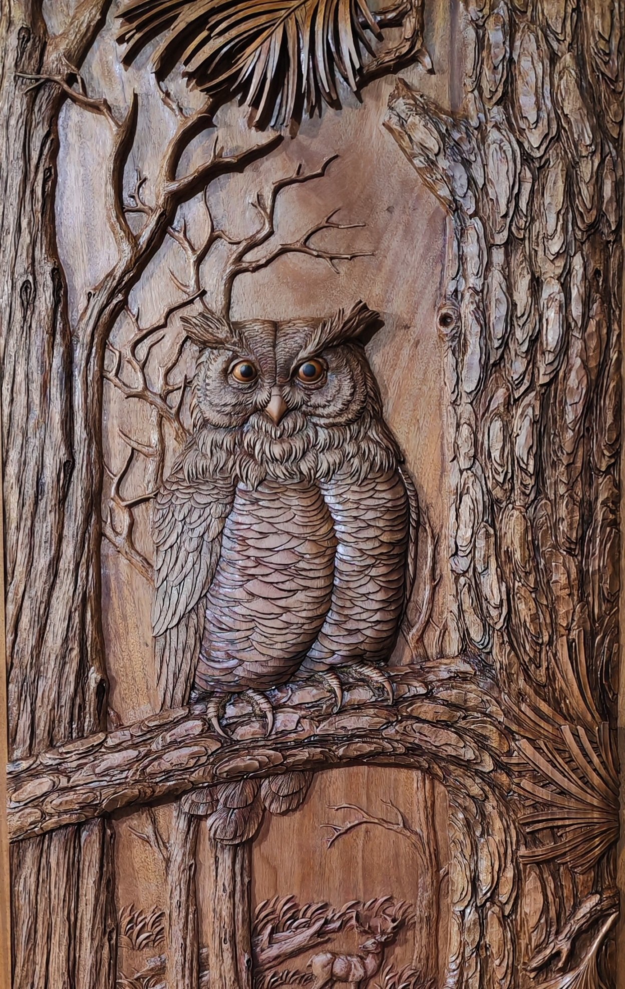 Close-up of a hand-carved horned owl surrounded by tree branches and textured bark on a wooden door