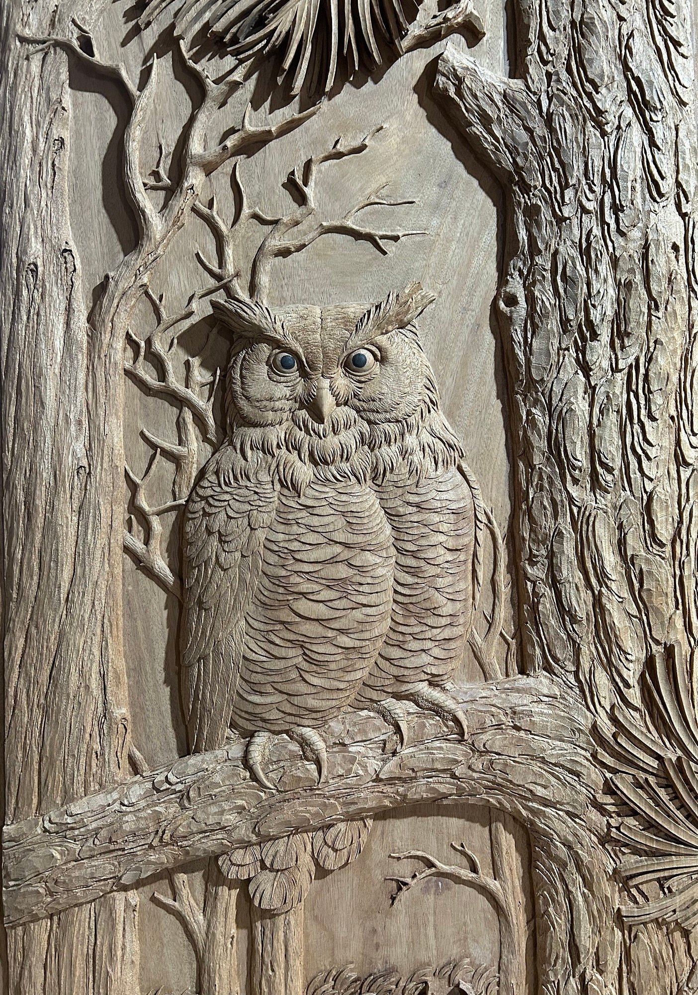 Detailed close-up photo of a deeply hand-carved horned owl with expressive blue inset eyes perched on a pine branch within a rustic forest relief setting on a solid wood door.